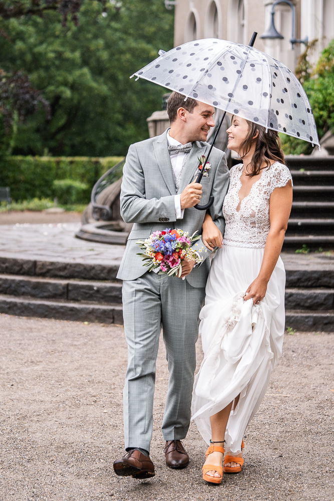 Foto von Lisa Werner Fotografie, Hochzeit, Brautpaar läuft lächelnd mit Regenschirm auf Fotografen zu