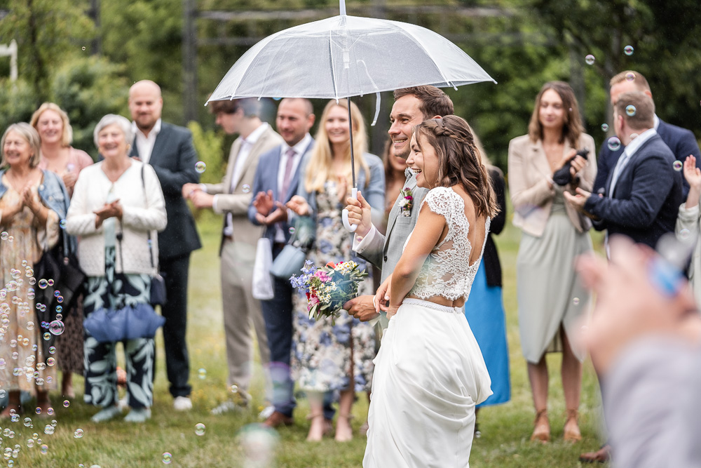 Foto von Lisa Werner Fotografie, Hochzeit, Brautpaar läuft lächelnd mit Regenschirm auf die Gäste zu