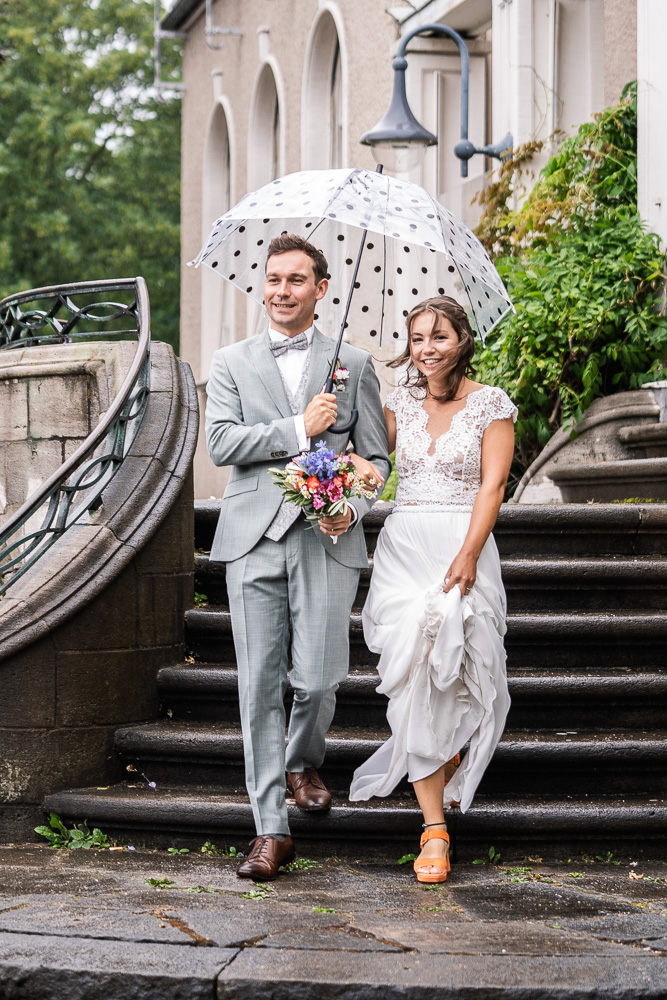 Foto von Lisa Werner Fotografie, Hochzeit, Brautpaar läuft lächelnd mit Regenschirm eine Treppe herunter
