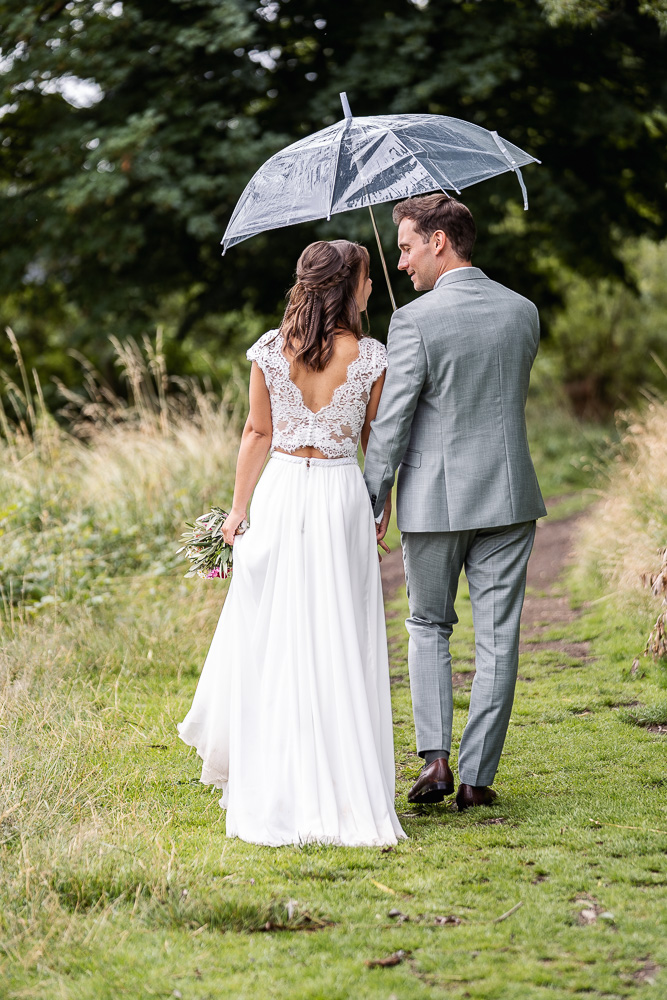 Foto von Lisa Werner Fotografie, Hochzeit, Brautpaarshooting, Brautpaar läuft mit Regenschirm über eine Wiese